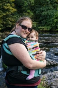 Helen cuddling her baby in a Tula carrier by a river