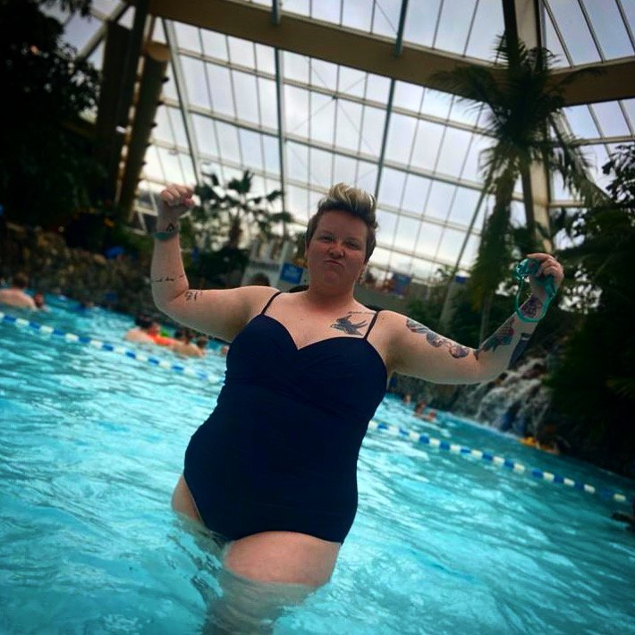 AJ is looking fierce in a black one-piece swimsuit in an indoor swimming pool, topped by a great glass roof and surrounded by palm trees and a small waterfall. Their tattooed arms are raised in a strong, defiant pose, with short blonde, punky hair and a face that says "bring it on!".