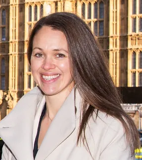 Image shows a smiling Dr Mairead Black, who has long brown hair and is wearing a cream coloured coat, stood in front of the Houses of Parliament.